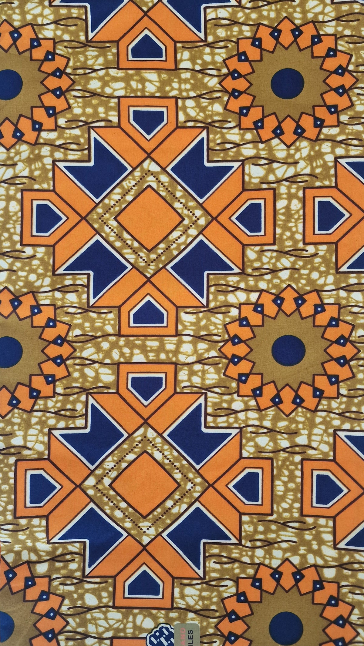 A folded piece of African wax print fabric with a geometric pattern in orange, navy blue, beige, and brown, featuring a batik-style design with circular medallions and starburst motifs.